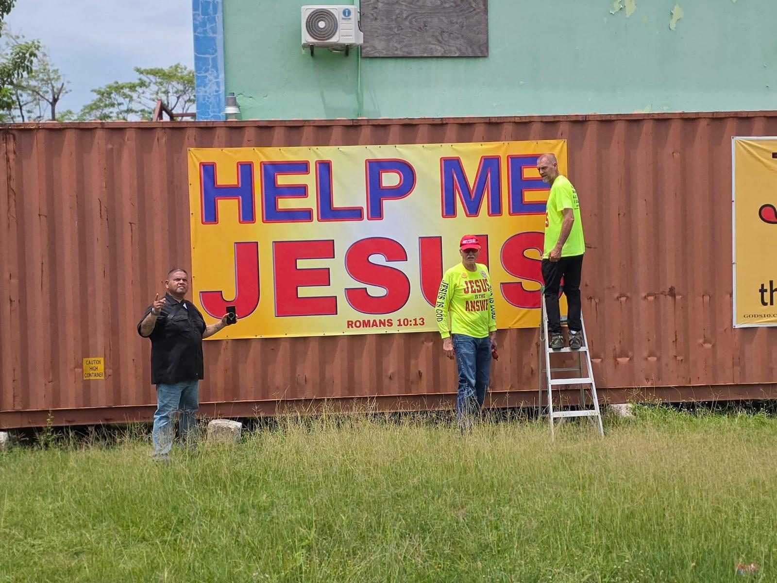A HELP ME JESUS banner, Romans 10:13, hung on a shipping container with volunteers nearby