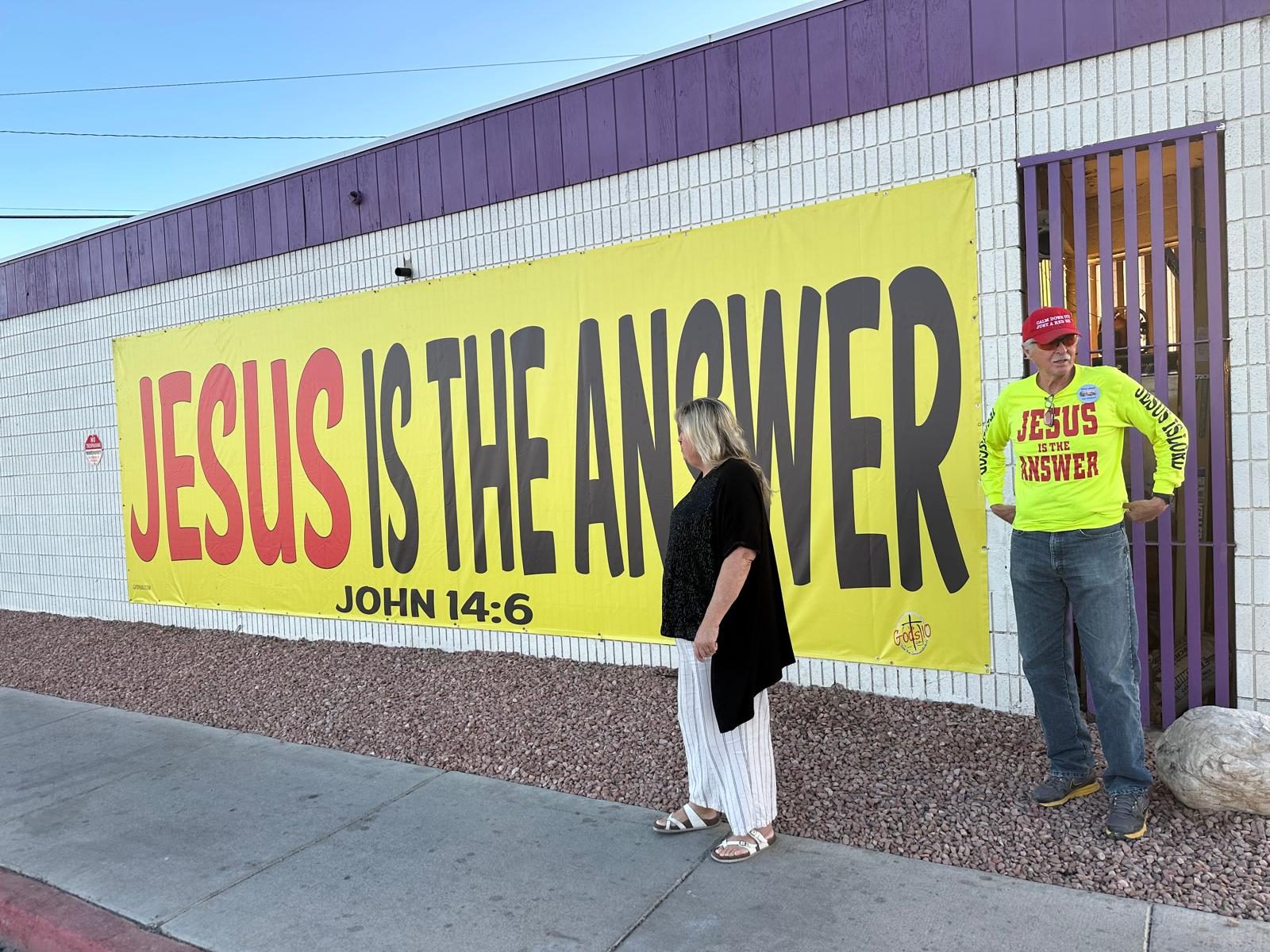 A JESUS IS THE ANSWER banner, John 14:6, along a city sidewalk wall