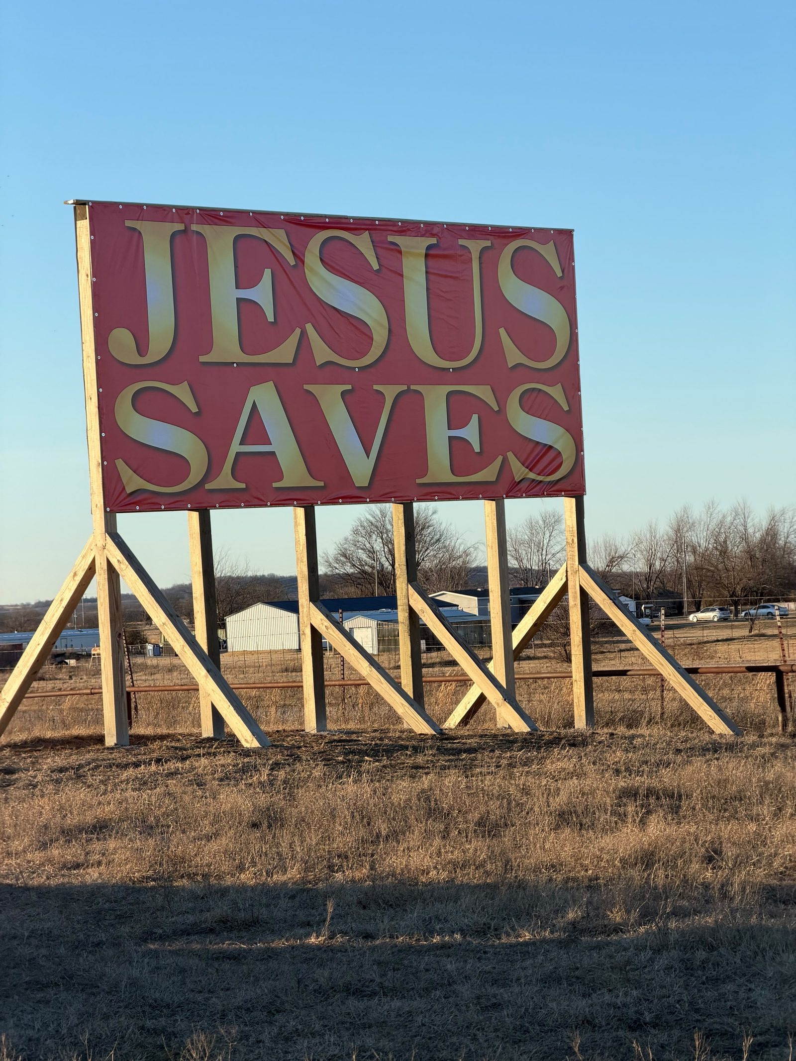A large JESUS SAVES banner in gold lettering on a red field, standing in an open field at golden hour