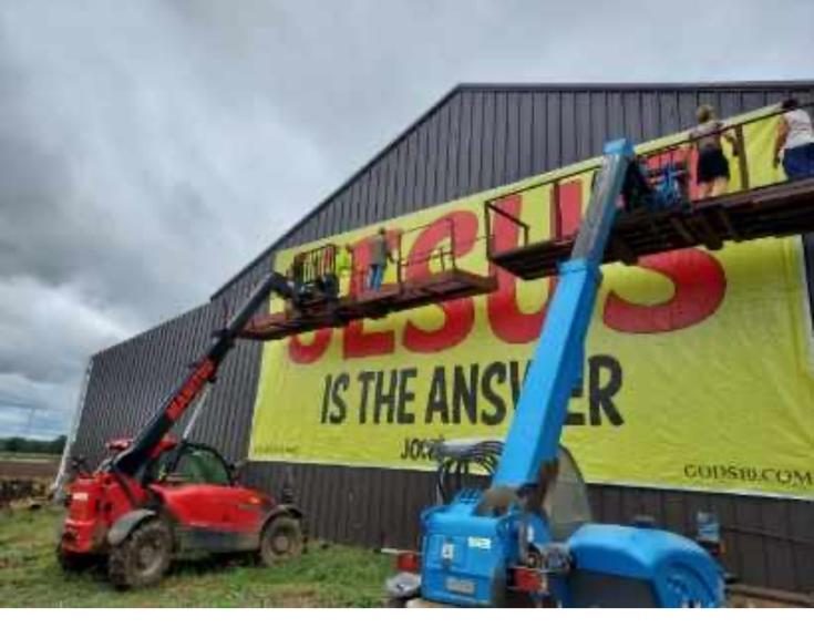 Two telehandlers and a crew raising a huge JESUS IS THE ANSWER banner on a building