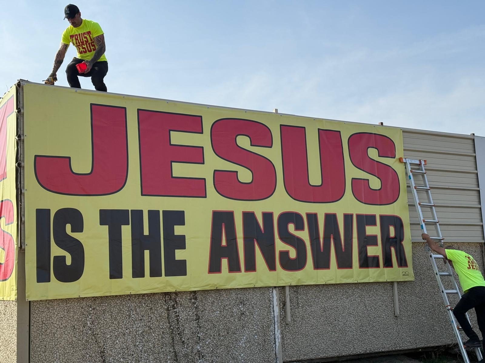 Volunteers in TRUST JESUS shirts securing a JESUS IS THE ANSWER banner from the rooftop and a ladder