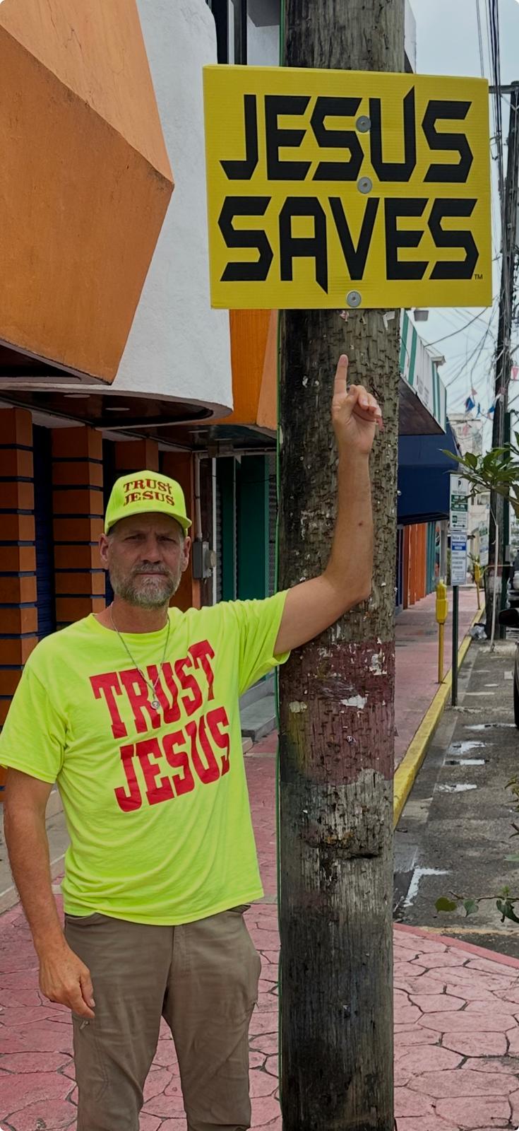 Steve Sawyer in a TRUST JESUS shirt pointing up at a JESUS SAVES sign on a street pole