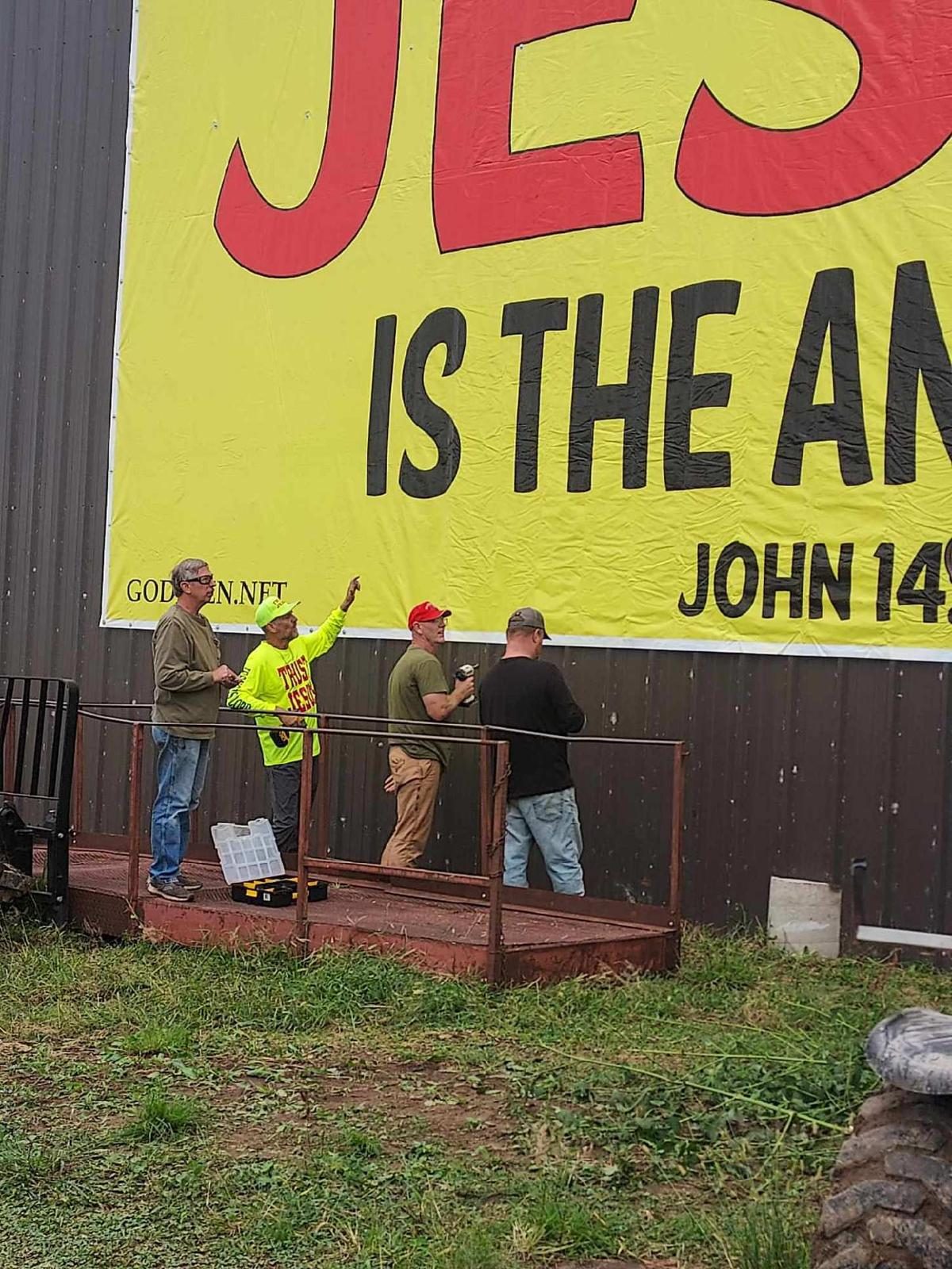 Volunteers gathered at the base of a freshly raised JESUS IS THE ANSWER banner